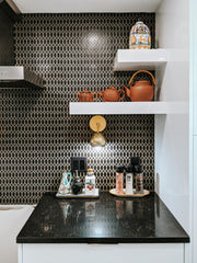 Black and white kitchen renovation with a brass mid century modern style sconce, black countertops, and black geometric tile backsplash with white floating shelves