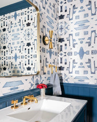 Modern glam bathroom renovation with a bold geometric blue and cream wallpaper, geometric black and brass wall sconces.  Bathroom features a white sink, brass hardware, and a large brass mirror with a natural linen washcloth.  Bathroom includes painted crown molding and beadboard wall trim.