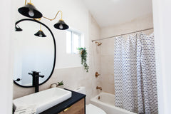 neutral bathroom renovation with a wood and black vanity with a black circular mirror and a black and brass wall sconce