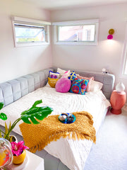 White bedroom with pops of color in musttard and bright pink.  Features a bright pink and brass wall sconce.  Used above a grey channel tufted day bed.