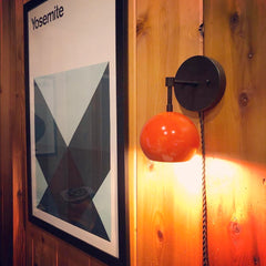 Black and orange mid century modern wall sconce on wood paneling and a black plug in cord. Has a 60s-70s style vibe with the graphic design Yosemite poster next to it.  Colorful playful space with a retro design style.