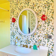Yellow floral wallpaper with red sconces, white mirror. In a bathroom with 50s style yellow bathtub tile.