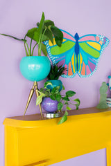 Maximalist living room color scheme featuring teal and brass large planter, a purple and chrome marbled planter, a mustard fieplace with rainbow butterfly wall art above it.