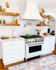 Modern kitchen with white tile walls, brass sconces, medium wood floating shelves, and white cabinets